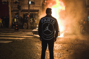 Man wearing a jacket with Michael Jordon logo on it. Fire is burning in front of him. In the background are some Gilet Jaunes protestors.